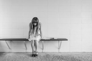 Eine Frau mit langem Haar sitzt auf einer Bank, die an an einer leeren Wand befestigt ist. Sie sitzt nach vorne gebeugt. Foto in Grautönen