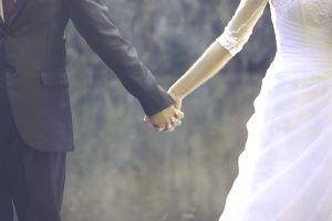 Links ein Bräutigam im Anzug, rechts eine Braut im Kleid halten einander an den Händen. Man sieht nur den Beginn ihrer Oberkörper und Teile der Beine.