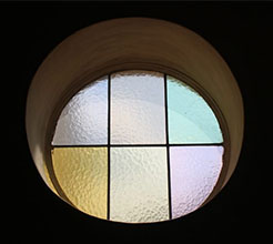 Ein rundes Glasfenster, mit Metallstangen in sechs Abschnitte unterteilt. Das Glas ist leicht gewellt und in blassen Pastellfarben: blau, rot, grün, gelb, blassgelb, rosa.