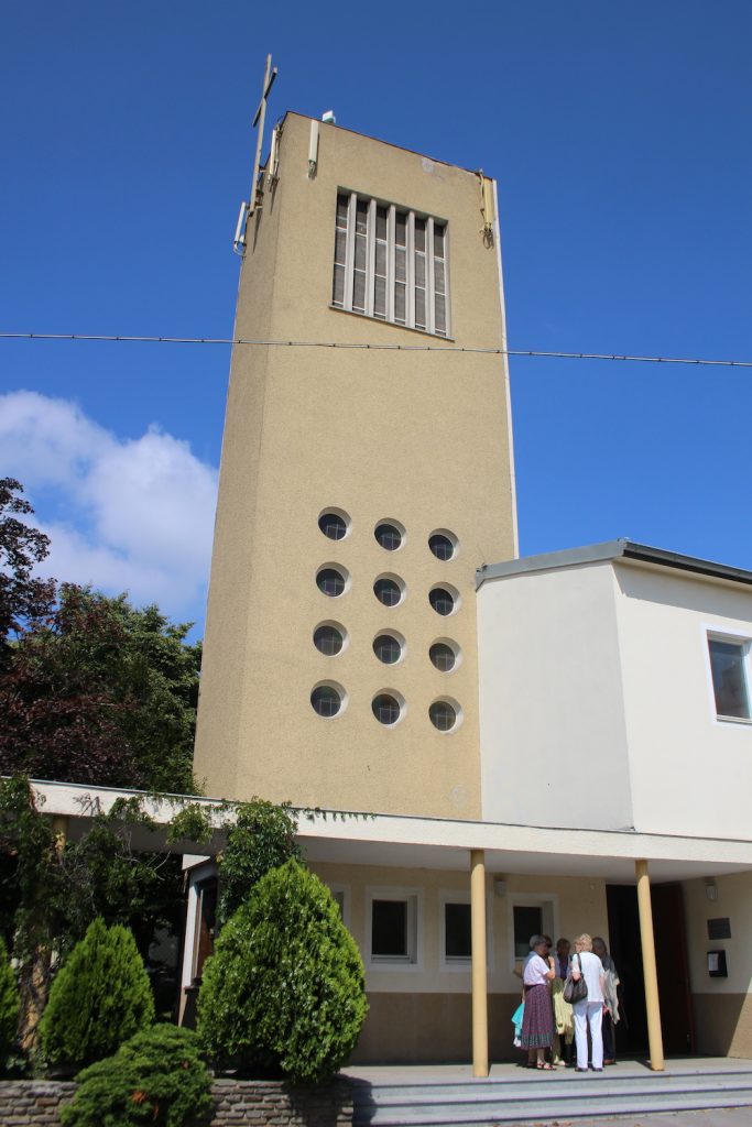 Foto: Hellgelber Kirchturm, oben flach mit Kreuz, weiter unten mit gitterartig angeordneten, kleinen runden Fenstern. Davor ein überdachter Säulengang.