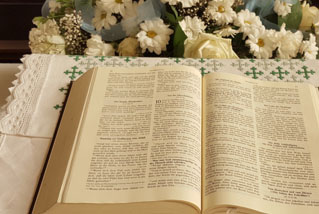 Eine aufgeschlagene Bibel in zweispaltigem Layout auf einer Zierdecke. Dahinter weißte Blumen.